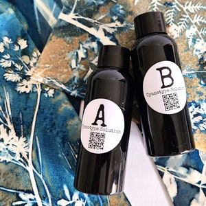 May include: Two black bottles labelled "A" and "B" with white circular stickers that read "Cyanotype Solution" and a QR code. The bottles are on a blue and brown floral patterned fabric.