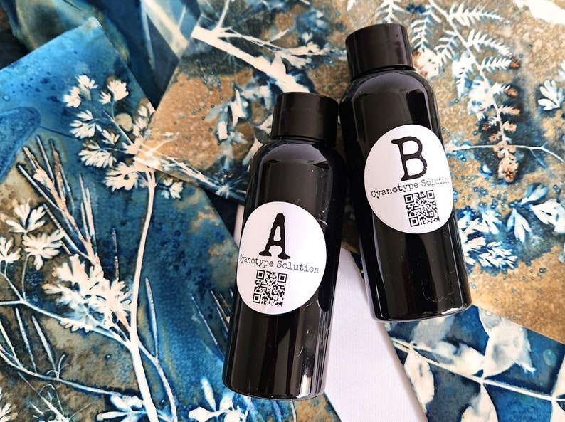 May include: Two black bottles labelled "A" and "B" with white circular stickers that read "Cyanotype Solution" and a QR code. The bottles are on a blue and brown floral patterned fabric.