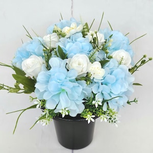 May include: A floral arrangement of artificial flowers in a black pot. The arrangement features light blue and white flowers, including large blue blooms, white roses, and small white accent flowers, with green foliage.
