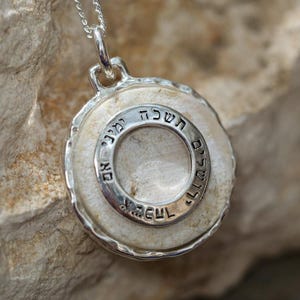 Może przedstawiać: Srebrny naszyjnik z zawieszką w kształcie białego kamienia z okręgiem z hebrajskim tekstem. Zawieszka jest na srebrnym łańcuszku.