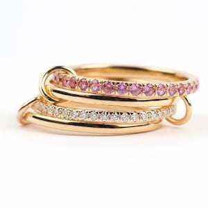 Könnte beinhalten: Ein Stapel goldener Ringe mit rosa und weißen Edelsteinen. Der oberste Ring hat eine Reihe rosafarbener Edelsteine, während der darunter liegende Ring eine Reihe weißer Edelsteine aufweist. Die Ringe sind miteinander verbunden und glänzend.