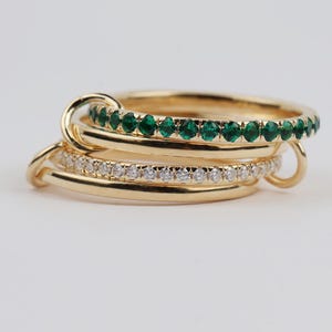 May include: A stack of four gold rings linked together. Two rings feature thin bands with embedded stones: one with vibrant green emerald-colored stones and another with clear white stones. The other two rings are plain, polished gold bands.