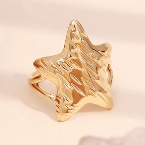 May include: A gold-toned ring with a textured star-shaped design. The ring is open on one side and has a smooth band.