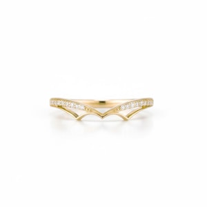 Puede incluir: Un anillo dorado con un diseño curvo, engastado con pequeñas piedras preciosas transparentes. El anillo tiene una forma única y puntiaguda en el centro y se muestra sobre un fondo blanco. Probablemente sea una alianza de boda o una joya de moda.