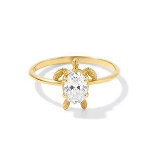 May include: A gold-colored ring featuring a turtle design. The turtle has textured details and holds an oval-shaped, clear gemstone. The ring band is thin and smooth, creating a delicate and elegant look.