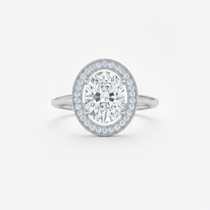 K&ouml;nnte beinhalten: Ein silberner Verlobungsring mit einem oval geschliffenen Diamanten als Mittelstein. Der Diamant ist von einem Halo aus kleineren Diamanten umgeben. Der Ring hat ein zartes Band und ist vor einem wei&szlig;en Hintergrund platziert.