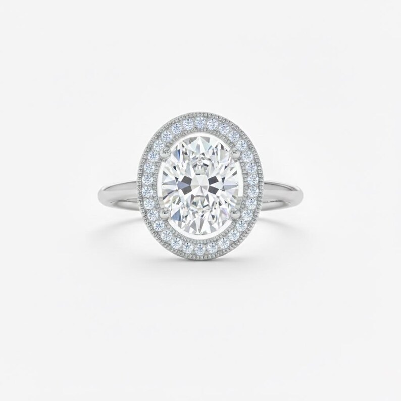 K&ouml;nnte beinhalten: Ein silberner Verlobungsring mit einem oval geschliffenen Diamanten als Mittelstein. Der Diamant ist von einem Halo aus kleineren Diamanten umgeben. Der Ring hat ein zartes Band und ist vor einem wei&szlig;en Hintergrund platziert.