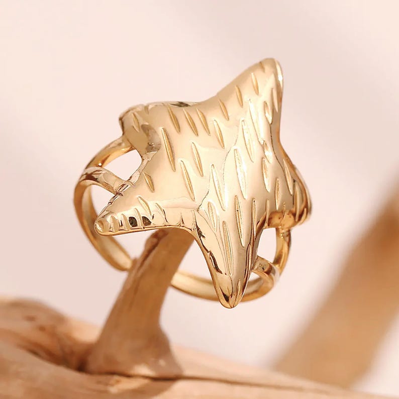 May include: A gold-toned ring with a star-shaped design. The star has a textured surface and is set in a simple band.