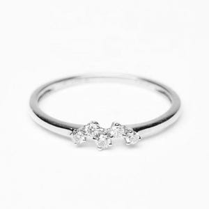 Puede incluir: Un delicado anillo de plata con un grupo de pequeñas piedras preciosas transparentes. El anillo tiene una banda fina y las piedras preciosas están dispuestas en forma curva, creando un diseño sutil y elegante. El anillo está sobre un fondo blanco liso.