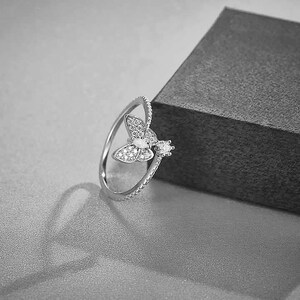 Peut inclure: Une bague argentée ornée d'un motif papillon. Le papillon est orné de petites pierres scintillantes, et le corps de la bague présente un motif texturé. Une seule pierre plus grande accentue le design. La bague est présentée sur une surface grise.
