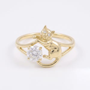 May include: A gold ring featuring a sculpted cat figure with two small clear stones for eyes. The cat is positioned next to a single, large, round, faceted clear stone held within the band, creating a playful and detailed jewellery piece.