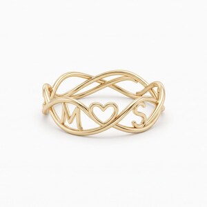 May include: A gold ring featuring an infinity-style twisted band design. The center of the ring displays the letter M, a heart shape, and the letter S, all integrated into the continuous metal structure of the band with a polished finish.