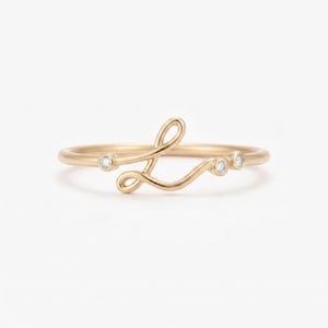 May include: A gold ring featuring a cursive letter L design as the centerpiece. The band is thin and polished, accented with three small, round, clear stones set in bezel settings along the flowing, script-like metal wire of the letter shape.