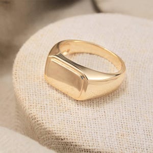 Könnte beinhalten: Ein goldener Siegelring mit rechteckiger Oberfläche. Der Ring hat eine polierte Oberfläche und liegt auf einer strukturierten Oberfläche in neutraler Farbe. Der Ring ist ein klassisches Schmuckstück.