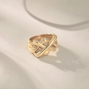 May include: A gold ring featuring a woven design. The ring has a thick band and a series of curved, overlapping elements, creating a textured appearance. The ring is set against a neutral background.