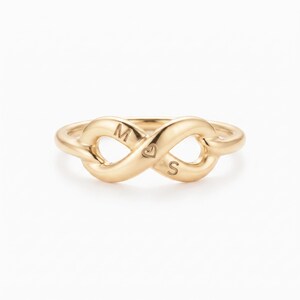 May include: A gold-toned ring featuring an infinity symbol design. The band is engraved with the letter M, a small heart icon, and the letter S, positioned along the centre of the infinity loop. The metal has a smooth, polished finish.