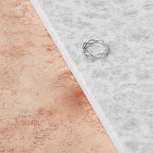 May include: A silver-colored ring with an interwoven design, set with small, clear gemstones. The ring is displayed on a white and gray marble surface, with a section of pinkish-brown stone in the background. The ring's intricate pattern is clearly visible.