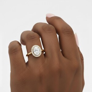 K&ouml;nnte beinhalten: Ein goldener Verlobungsring mit einem oval geschliffenen Diamanten, umgeben von einem Halo aus kleineren Diamanten. Der Ring ist auf einem zarten Goldband gefasst. Der Ring wird an einem Finger gezeigt.