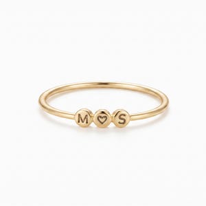 May include: A thin gold ring featuring three small, circular charms centred on the band. The left charm is engraved with the letter M, the middle charm displays a heart symbol, and the right charm is engraved with the letter S.