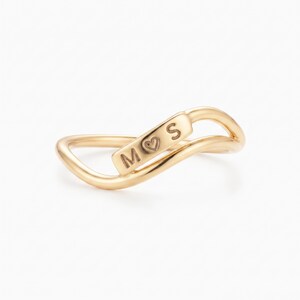 May include: A gold-toned ring featuring a wavy, open-band design. A central rectangular plaque is integrated into the band, engraved with the letter M, a heart symbol, and the letter S. The metal surface has a smooth, polished finish throughout.