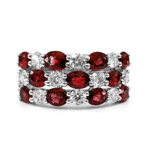 Pode incluir: Anel de três fileiras com pedras vermelhas ovais alternadas com pedras claras redondas, cravejadas em uma banda prateada. O anel é exibido em um fundo branco.