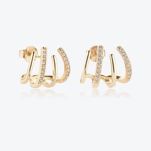 May include: A pair of gold-coloured earrings. Each earring has a three-pronged design, with one prong set with small, clear gemstones. The earrings have a post-back closure. The jewellery is displayed on a white background.
