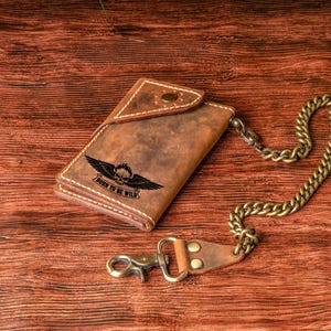 May include: Brown leather wallet with a chain, featuring a skull and wings graphic with the text "BORN TO BE WILD". The wallet has a snap closure and a metal chain with a clasp.