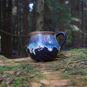 May include: A handmade ceramic mug with a dark blue, brown, and white glaze. The mug has a black handle and sits on a wooden surface. The mug is approximately 4 inches tall.