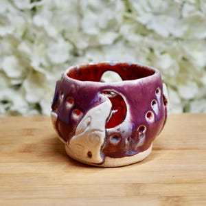 Könnte beinhalten: Ein handgefertigter Keramik-Kerzenhalter mit Wolfsmotiv. Der Halter ist tiefviolett mit rotem Innenraum und weißen Wolfsdetails. Er hat mehrere kleine Löcher und steht auf einer Holzoberfläche.