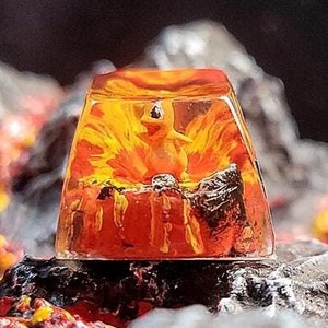 May include: A clear, rectangular resin art piece with a fiery orange and yellow interior, featuring a small, yellow figure. The piece sits atop a dark, textured surface with a blurred background of gray and red elements, evoking a volcanic theme.
