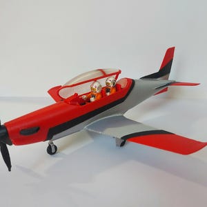 Peut inclure: Un avion jouet rouge et gris avec une verrière transparente. Deux pilotes jouets sont assis dans le cockpit. L'avion a une hélice noire et un train d'atterrissage.