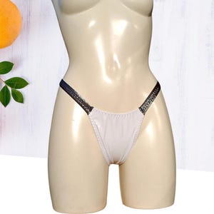 Puede incluir: Braga tanga blanca con acabado brillante y tirantes gris oscuro. La ropa interior se exhibe en un maniquí. El fondo es una superficie de madera blanca con una vista parcial de una fruta y hojas.
