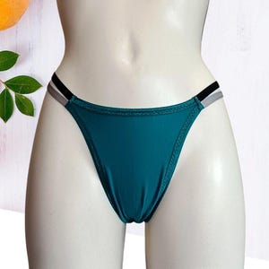 Puede incluir: Braguita de bikini verde azulado con tirantes negros y grises. La ropa de baño se exhibe en un maniquí. La braguita tiene un diseño de corte alto en la pierna y una textura suave y brillante. El fondo incluye una superficie blanca y una vista parcial de una fruta.