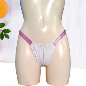 Puede incluir: Ropa interior de satén blanco con ribete lavanda. La ropa interior tiene una cintura baja y un corte atrevido.