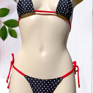 Op de afbeelding: Zwart-witte bikini met witte stippen en rode bandjes. De top is van het driehoekstype en de onderkant is van het ondeugende type.