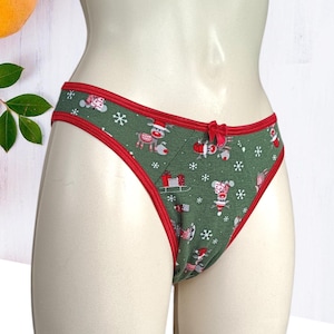 Puede incluir: Braga verde con ribete rojo y un pequeño lazo rojo. La ropa interior presenta un estampado navideño con renos, copos de nieve y cajas de regalo. La ropa interior se exhibe en un maniquí.