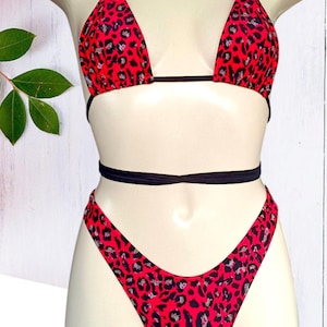 Könnte beinhalten: Rotes und schwarzes Bikini-Set mit Leopardenmuster. Das Triangel-Oberteil hat schwarze Träger, und die Unterteile sind hoch geschnitten. Der Bikini wird auf einer Schaufensterpuppe vor weißem Hintergrund präsentiert.