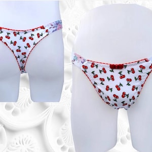 Peut inclure: String blanc avec un motif de cerises. Les bords sont bordés de coutures rouges et la ceinture présente un motif floral. Un petit nœud rouge est centré sur le devant.