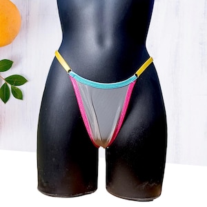 Puede incluir: Un tanga transparente exhibido en un maniquí negro. El bikini presenta una tela de malla gris con ribetes rosas y turquesas. Las correas amarillas se conectan a los lados. El maniquí está colocado sobre un fondo blanco.