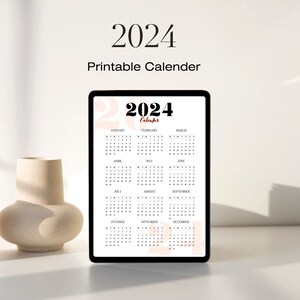 May include: A printable 2024 calendar on a tablet screen. The calendar is in black and white with red text for the months and holidays. The background is a light pink color.