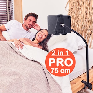 Puede incluir: Un soporte de teléfono o tableta negro con un brazo largo y flexible, sujeto a una abrazadera. El soporte está colocado sobre una cama con dos personas acostadas en ella. El texto "2 en 1 PRO 75 cm" es visible en un círculo rojo.