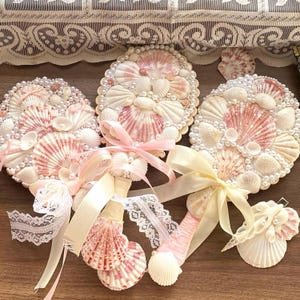 Op de afbeelding: Decoratieve kunstwerken van zeeschelpen. De stukken zijn rond en tonen witte en roze zeeschelpen, gerangschikt in ingewikkelde patronen, geaccentueerd met parels. Elk stuk is versierd met linten en kant. De achtergrond is een houten oppervlak.