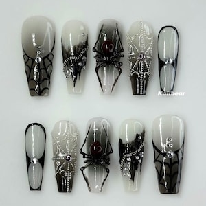 Könnte beinhalten: Ein Set aus zehn künstlichen Nägeln mit Halloween-Motiv. Die Nägel weisen einen Farbverlauf von Weiß zu Schwarz, Spinnennetze, Spinnen und dekorative Ketten auf. Einige Nägel haben Strasssteine. Der Markenname "Nailbear" ist auf einem Nagel sichtbar.