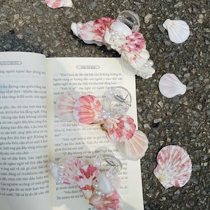 Puede incluir: Tres pinzas para el pelo con forma de concha marina con cierres de plástico transparente. Las pinzas presentan un estampado floral rosa y blanco, adornado con perlas de imitación. Las pinzas están sobre un libro abierto.