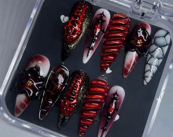 May include: A set of ten long, pointed false nails in a clear plastic case. The nails feature a variety of designs, including textured red, black and gold glitter, and dark red and white gradients. Some nails have floral or cross-like embellishments.