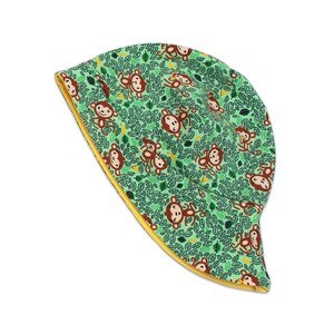 Può includere: Un cappello da sole verde e giallo con un motivo ripetuto di scimmie marroni e foglie verdi. Il cappello è in tessuto e ha un bordo giallo.