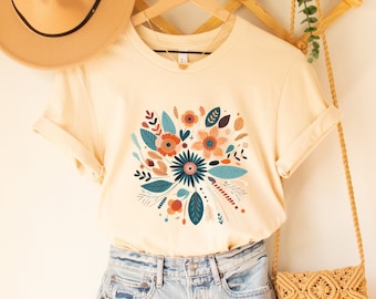 May include: A cream colored t-shirt with a floral design in shades of blue, orange, and brown. The shirt is on a hanger and has short sleeves.