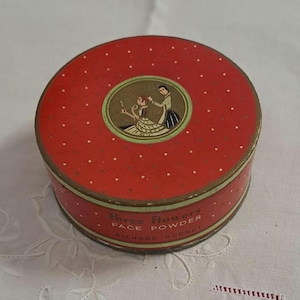 Könnte beinhalten: Eine runde Blechdose mit einem roten Deckel mit weißen Punkten. Der Deckel zeigt eine Vintage-Illustration von zwei Personen. Der Text auf der Unterseite lautet "Three Flowers Face Powder Richard Hudnut".