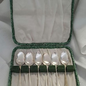Könnte beinhalten: Sechs silberne Löffel in einem geöffneten, grün-weiß gefütterten Etui. Die Löffel haben ein klassisches Design mit einem dekorativen Muster am Boden der Laffe. Das Etui hat einen grünen, gemusterten Rand und einen Verschluss.