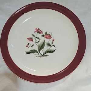 Op de afbeelding: Een wit keramiek dinerbord met een bordeauxrode rand. Het midden van het bord heeft een bloemmotief met roze bloemen en groene bladeren.